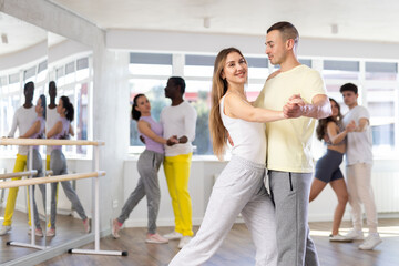 Positive young adult couple in casual sportswear enjoying slow foxtrot in sunny dance studio....
