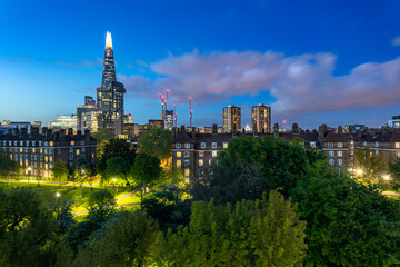 Fototapeta na wymiar A residental neighbourhood with a geen park and illuminated buildings including skyscrapper Shard in London during blue hour