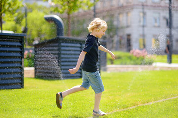 Funny little boy playing with lawn sprinkler in sunny city park. Elementary school child laughing, jumping and having fun with spray of water. - 791185544