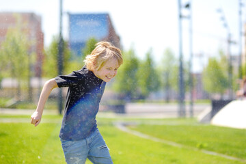 Funny little boy playing with lawn sprinkler in sunny city park. Elementary school child laughing, jumping and having fun with spray of water. - 791185705