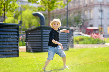 Funny little boy playing with lawn sprinkler in sunny city park. Elementary school child laughing, jumping and having fun with spray of water. - 791185932