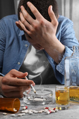 Alcohol and drug addiction. Man with smoldering cigarettes, whiskey and pills at grey textured table - obrazy, fototapety, plakaty