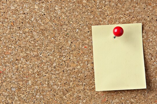 A rectangular post-it stuck delicately against a wooden cork board in a simple, familiar image. A post-it contrasts with the natural tone of the cork as a highlight on the textured surface.