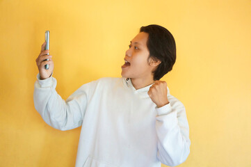 Excited Asian young man wearing a hoodie while holding and looking at smartphone
