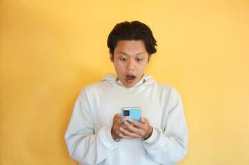 Fototapeta premium Surprised Asian young man wearing a hoodie while holding and looking at mobile phone