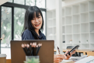 Businesswoman smiles while holding a smartphone and writing notes, laptop open and stationery on desk, in a bright office setting.