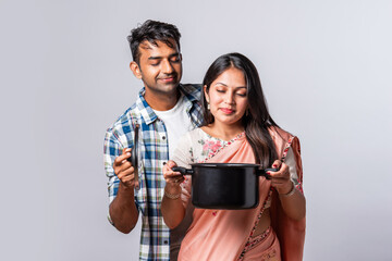 Asian indian young couple standing with kitchen utensils against plain background