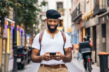 Stylish black man walking on the city street using smartphone