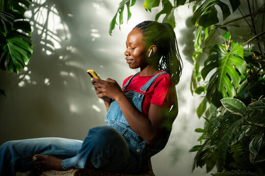 Relaxed African American woman wear earphones looks at smartphone screen, engrossed in selects music track on mobile app. Black girl holds phone scrolling social media at home garden. Slow life, rest.