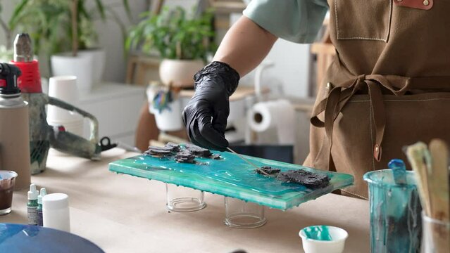 Female artist pours epoxy resin and smears it with stick on wooden board. Craftswoman using liquid art technique, creating picture of sea and waves, rocks closeup view. Master class on resin art. 