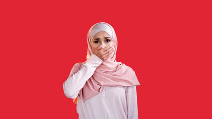 Secret fear. Shocked face. Dismay panic. Scared disturbed upset woman in headscarf covering mouth with hand isolated on red empty space background.