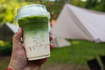 Cropped hand holding iced green tea matcha latte with camping background