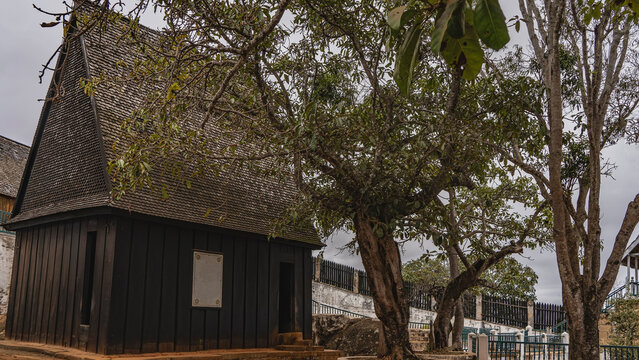 The complex of the ancient royal palace. A dark wooden house Mahandrihono with a triangular tiled roof. Sprawling trees. The fence is far away. Madagascar. Antananarivo. Royal Hill of Ambohimanga