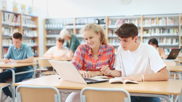 Portrait of two focused schoolchildren doing homework in school library, sitting at desk in front of laptop
