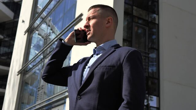 Businessman swiftly raises phone to ear backdrop of office building. Businessman engages in serious dialogue, intensity marked. Businessmans strategic conversation underscores his focus and expertise.