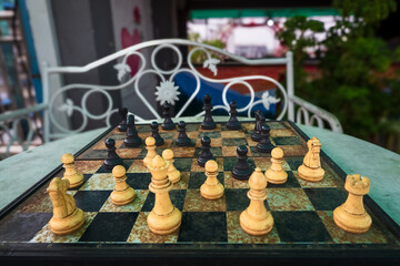 Incomplete set of chess pieces arranged on a rusted chess board with a bench in the background