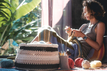 A woman sits on the floor and knits a multicolored blanket from woolen threads in the foreground is...