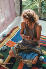 A woman sits on the floor in the bedroom and knits a blanket from woolen threads. The background in...