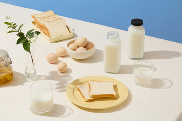 Preparation for cooking a meal with a set of ingredients including egg, milk, sandwich decorated by a flower vase on white table, blue background. Top view, blank space