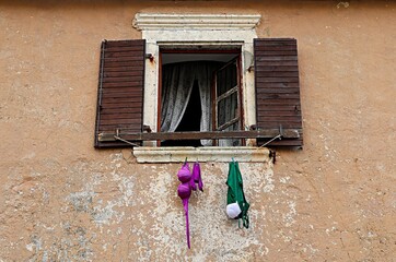 An open window with framed shutters; under the window, multi-colored items of a woman’s swimsuit are dried on a rope.