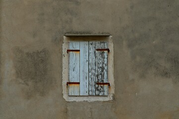 The window of an old house is closed with wooden shutters on rusty hinges, covered with white paint peeling off from age.