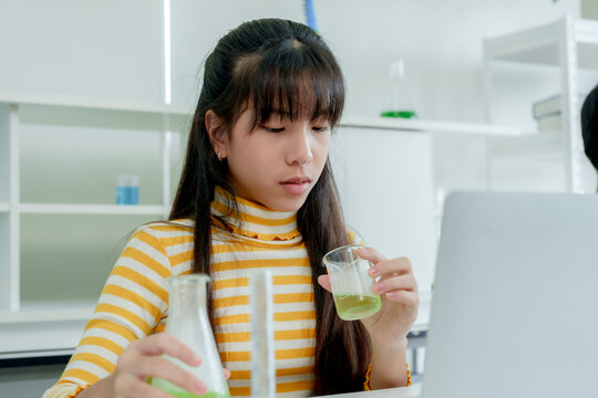 Students' learning in science classroom. Students in casual attire collaborate on science project, examining green chemical in flask, microscope and laptop on desk in bright classroom.