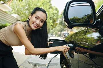 Happy woman inserting plug in electric car to charge battery