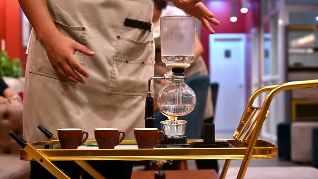 Close-up of a barista preparing coffee using a syphon brewer on a cafe table, with customer in
