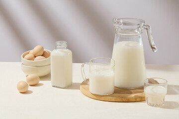 Different types of glass containers filled with fresh milk are arranged on white surface. A stack of ceramic bowls containing eggs. Tasty healthy dairy products on a table