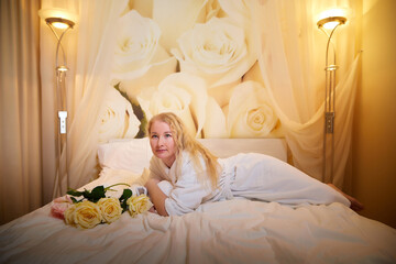 A blonde girl in a white terry bathrobe in a hotel room with flowers. Wedding travel, Weekend and vacation, body care