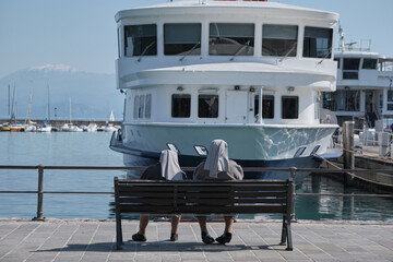 two nuns with gray headscarves are sitting on a bench opposite the ship, rear view