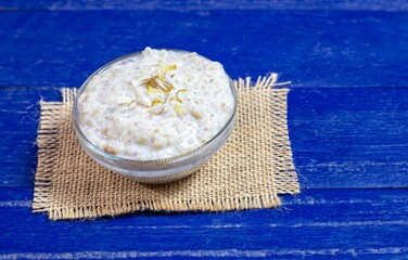 Cracked Wheat Kheer or Payesh Garnished with Cashew, Pistachio, Almond in a Glass Bowl Isolated on Blue Wooden Background with Copy Space, Also Known as Ksheeram, Milk Pudding