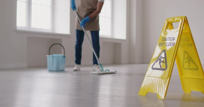 Close-up shot of a Caution, Wet Floor sign with a male cleaner mopping the floor in the background. This warning sign is a safety precaution to prevent slips and falls during cleaning.
