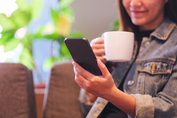 Closeup image of a young woman holding and using mobile phone while drinking coffee in cafe - 791325323