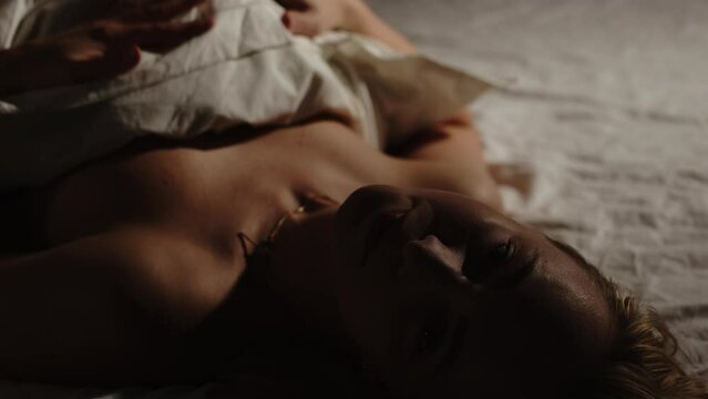 A beautiful young woman lays in a dark bedroom while hugging a pillow and smiling at camera