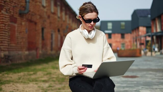 Serious woman holding laptop while sitting in urban place and typing numbers from credit card. Female in sunglasses using digital device for controlling financial transactions in bank application.
