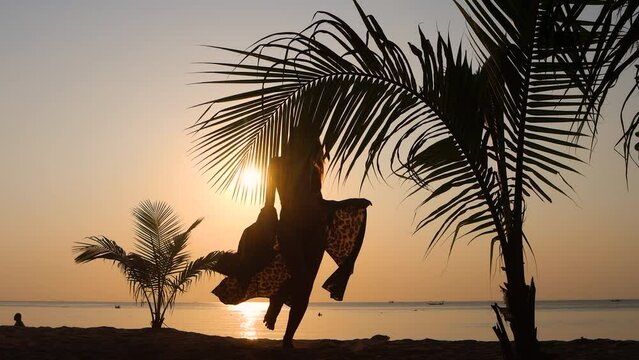 Young beautiful caucasian woman dancing on tropical sunset sea beach under palm leaves. Girl in leopard dress on vacation feel happy.