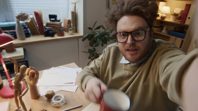 Medium high-angle UGC selfie shot of funny male corporate worker with curly red hair, in glasses sitting at desk, filming himself working on report for social media, toasting with mug of coffeeMedium 