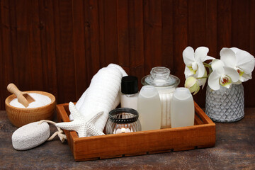 Spa arrangement with body care products, bath additives, aroma oil, hair shampoo, soap and towel in front of a wooden wall with space for text.