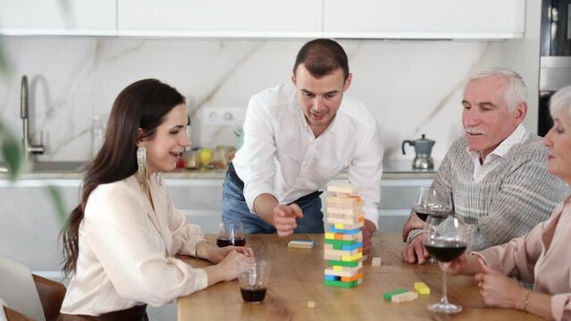 Young man playing jenga with family in cozy home kitchen, trying to pull out block without destroying tower, under interested glances of wife and elderly parents sitting at table with glasses of wine