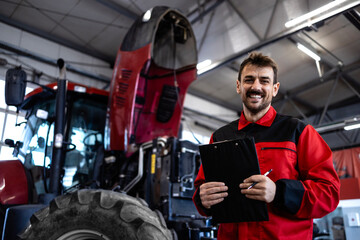 Portrait of smiling serviceman holding checklist by the tractor inside workshop.