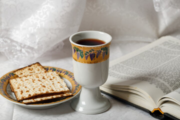 Kosher wine cup and matzah bread placed beside an open book. The backdrop is an elegant white fabric. Cup is adorned with intricate designs and the matzah rests on a decorative plate. Jewish pesach