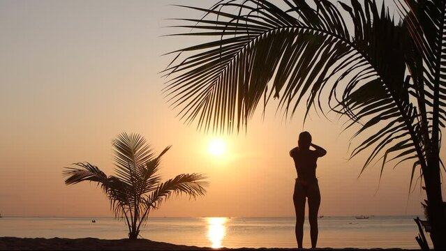 Young beautiful caucasian woman make yoga breath exercise on tropical sunset sea beach under palm leaves.