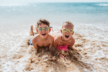 Siblings playing on beach, lying in water, having fun. Smilling girl and boy in swimsuits, swimming googles on sandy beach of Canary islands. Concept of family beach summer vacation with kids. - obrazy, fototapety, plakaty