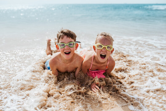 Siblings playing on beach, lying in water, having fun. Smilling girl and boy in swimsuits, swimming googles on sandy beach of Canary islands. Concept of family beach summer vacation with kids.