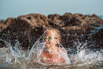 Young girl playing, swiming and splashing in fresh sea water. Smilling blonde girl in swimsuit with and swimming goggles. Concept of beach summer vacation with kids.