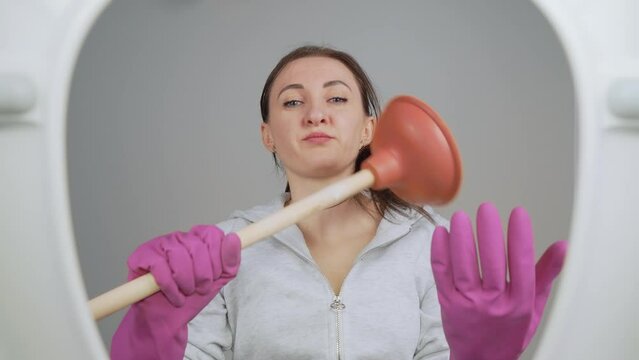 Focused woman stands ready to tackle plumbing issues as she confidently holds plunger in hands. Female wearing purple rubber gloves prepared for toilet cleaning. View from toilet bottom. Cleanliness