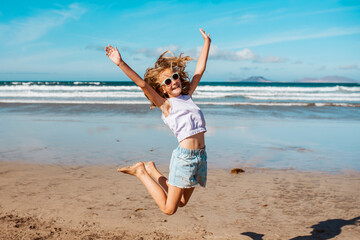 Jumping girl on beach. Smilling blonde girl enjoying sandy beach, looking at crystalline sea in Canary Islands. Concept of beach summer vacation with kids. - obrazy, fototapety, plakaty