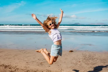 Fotobehang Jumping girl on beach. Smilling blonde girl enjoying sandy beach, looking at crystalline sea in Canary Islands. Concept of beach summer vacation with kids. © Halfpoint