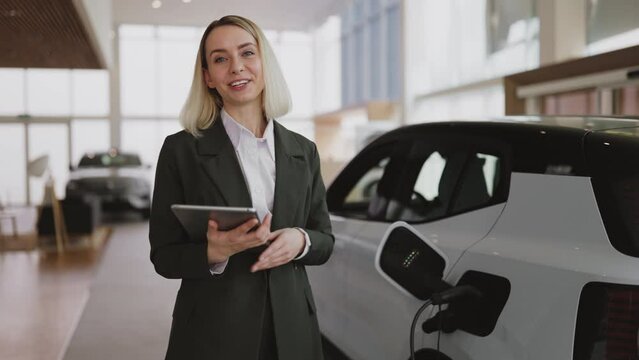 Young blond Caucasian saleswoman, selling electric cars in light modern showroom with panoramic windows. Concept of buying eco-friendly car for progressive customer.